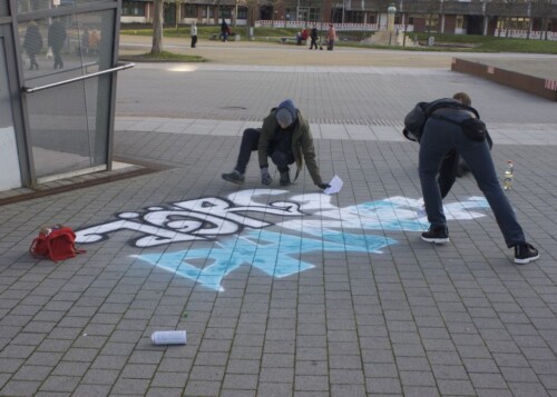Zwei Menschen sprühen den Namen von Jörg Danek auf den Boden vor dem Bahnhof